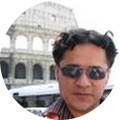man with short, dark hair wearing sunglasses and standing in front of the roman colliseum